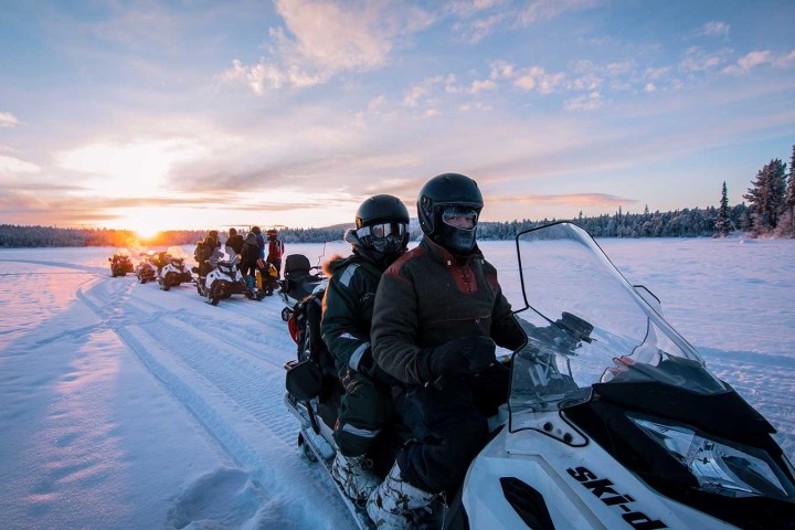 People riding snowmobiles on a snowy trail at sunset with trees in the background.