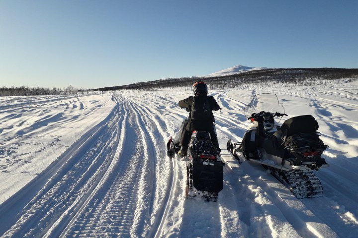 Person on a snowmobile beside another in a snowy landscape with mountains and clear sky.