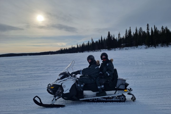 Two people on a snowmobile wearing winter gear, with a snowy landscape and trees in the background.