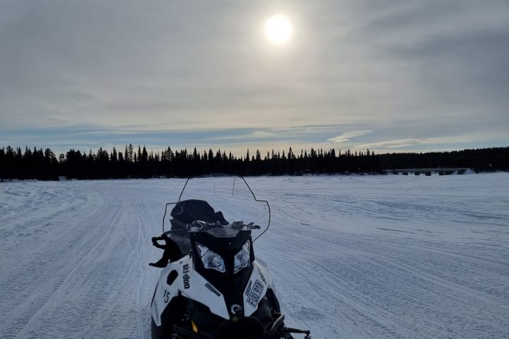 Snowmobile on snowy terrain beneath cloudy sky with sun overhead.