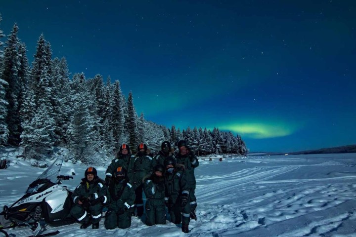 People in winter gear with snowmobiles under northern lights and snowy trees at night.