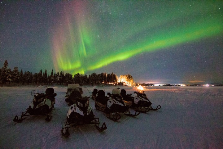 Snowmobiles parked on snow under a green aurora in a starry night sky.