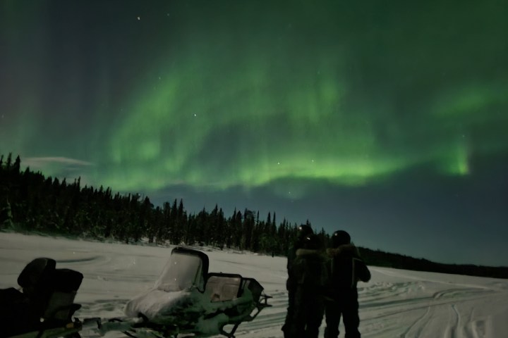 Two people and snowmobiles beneath green aurora borealis in snowy landscape.