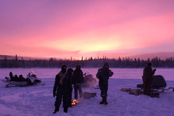 People by a small fire in snowy landscape with snowmobiles and a vibrant pink sky.