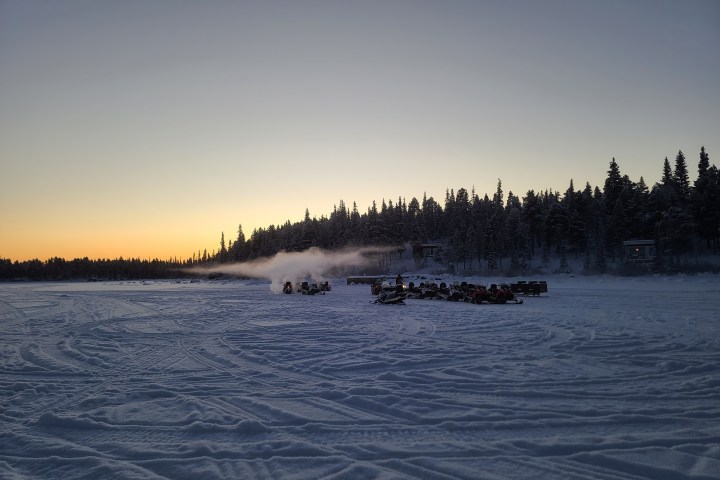 Sunset over snowy landscape with trees and a group of snowmobiles.
