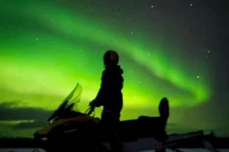 Silhouette of a person on a snowmobile with green aurora borealis in the night sky.