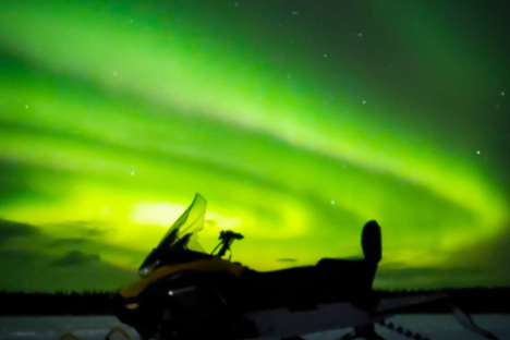 Snowmobile on snowy ground silhouetted against vibrant green aurora borealis in night sky.