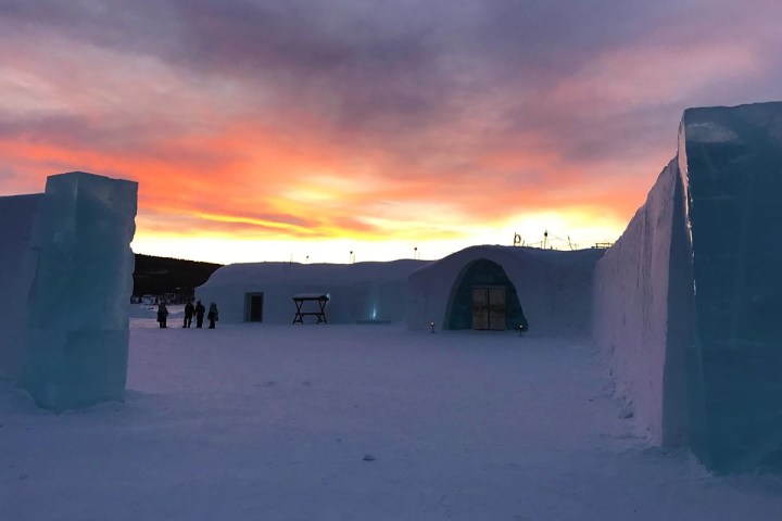Ice hotel buildings at sunset with vibrant sky and distant silhouettes of people.
