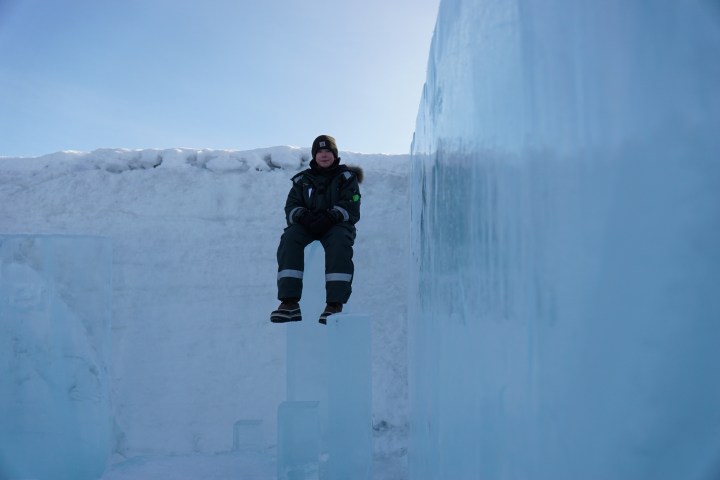 Person in winter clothing sitting on an ice block against a snowy, icy backdrop.