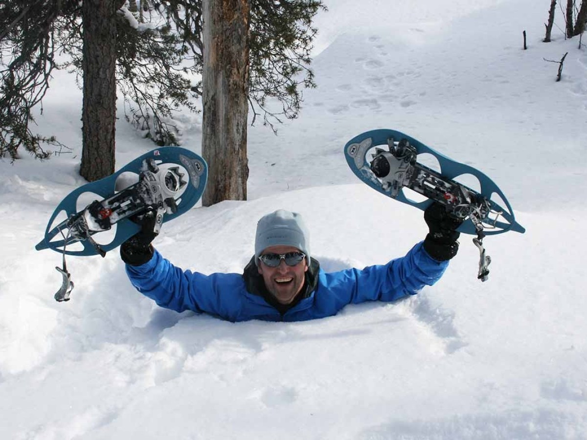 Person emerging from deep snow holding a snowshoe in each hand, wearing a blue jacket and sunglasses.