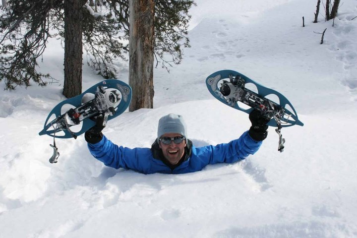 Person emerging from deep snow holding a snowshoe in each hand, wearing a blue jacket and sunglasses.
