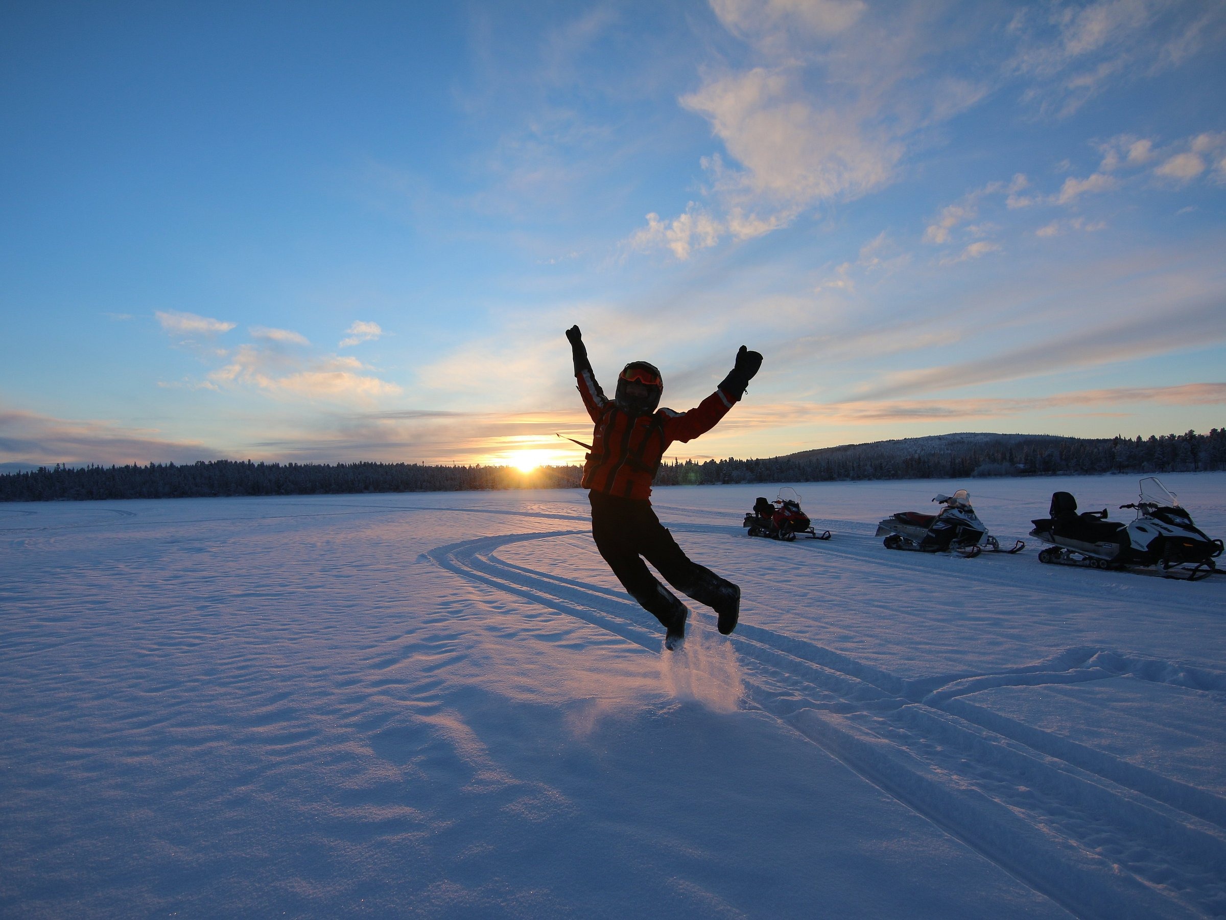 Person in winter gear jumps on snow with sunset and snowmobiles in background.