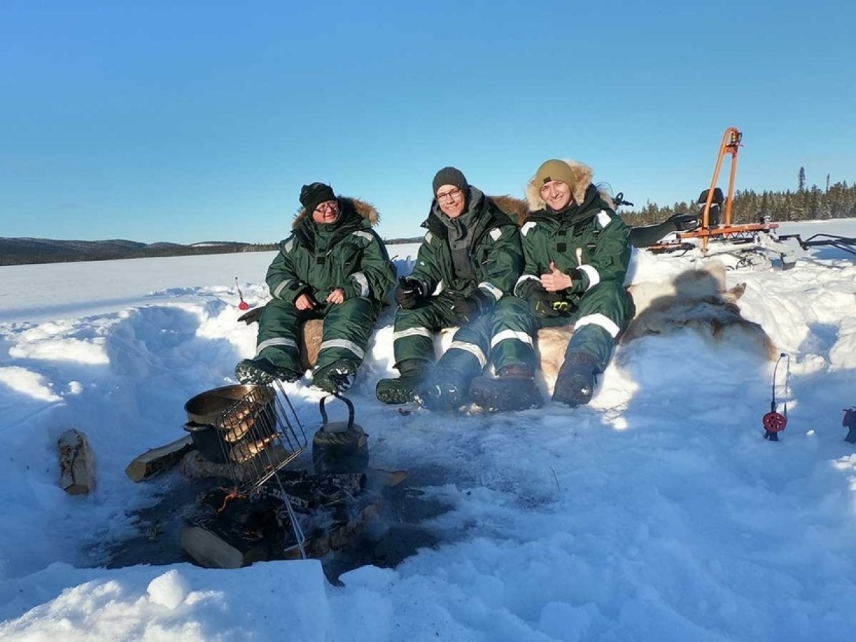 Three people in green winter suits sitting on snow near a campfire with a snowy landscape in the background.