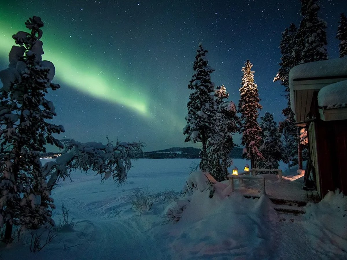 Snowy landscape with northern lights over trees and a cabin.
