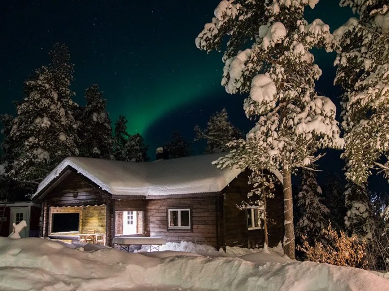 Cozy cabin in snowy forest with Northern Lights in sky above.