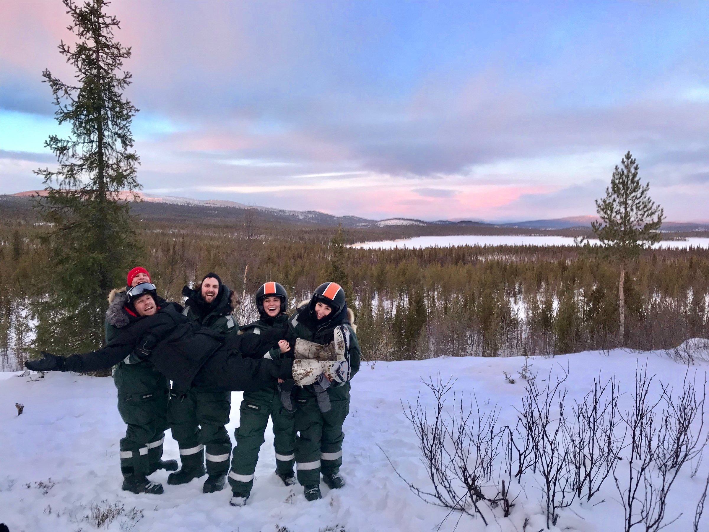 Group of people in winter gear posing with snowy forest and mountain landscape at sunrise.