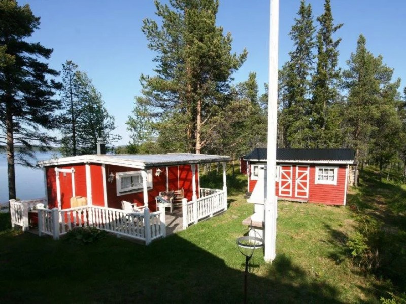Two red cabins in a forested area near a lake under a clear blue sky.