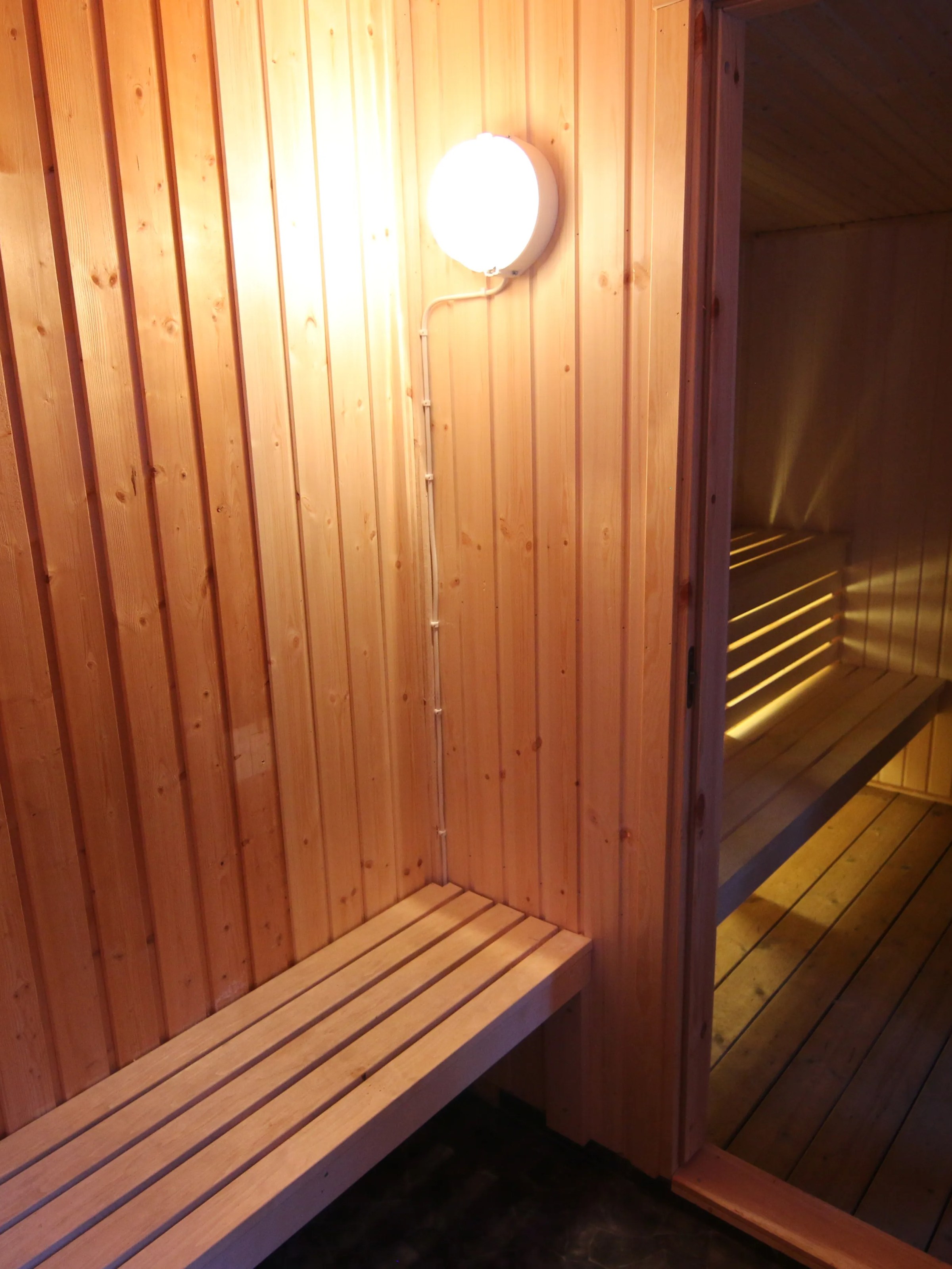 Wooden sauna with benches, a door, and a lit lamp on the wall.