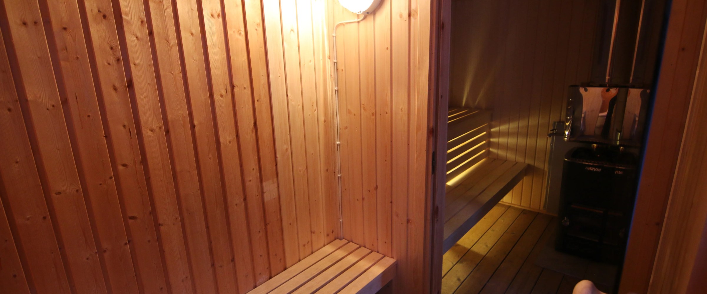 Wooden sauna with benches, a door, and a lit lamp on the wall.