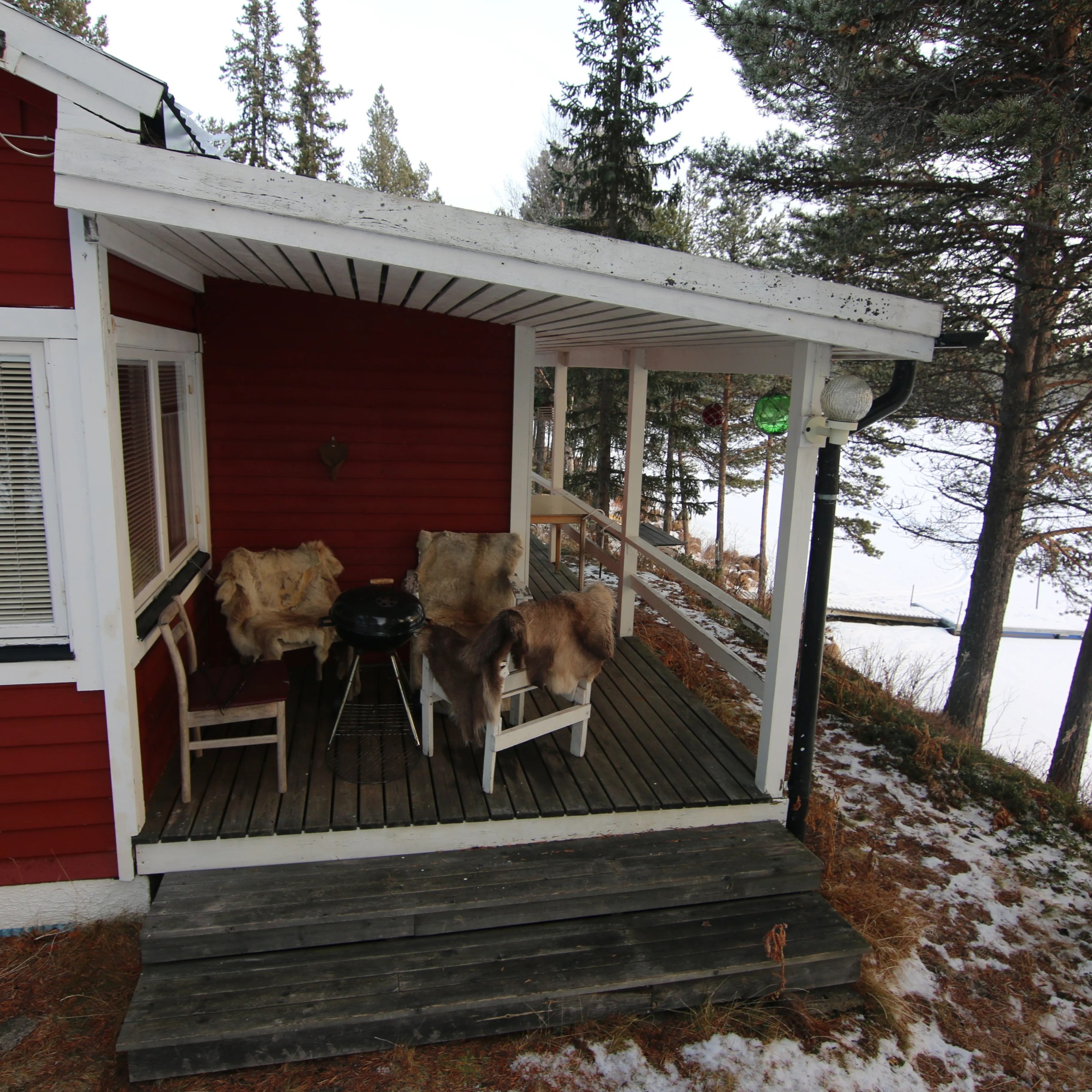 Red cabin porch with chairs and table, overlooking snowy woodland and frozen lake.