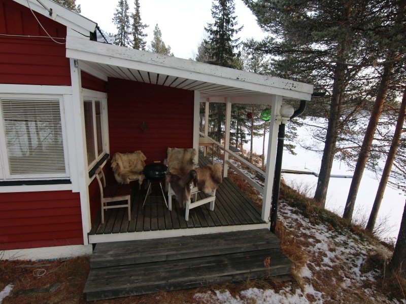 Red cabin porch with chairs and table, overlooking snowy woodland and frozen lake.