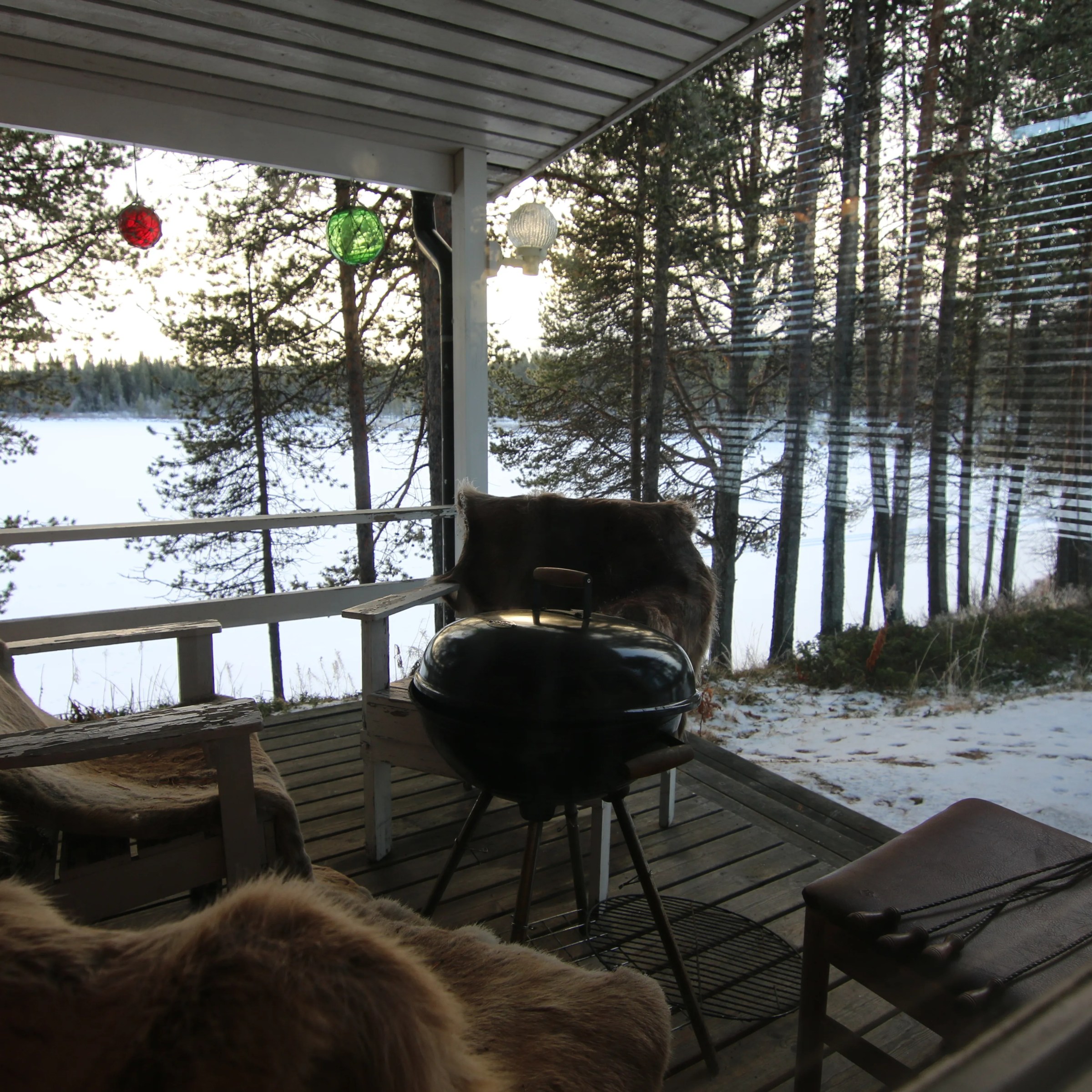 Snowy porch with fur-covered chairs, BBQ grill, and hanging colorful lights overlooking a lake and trees.