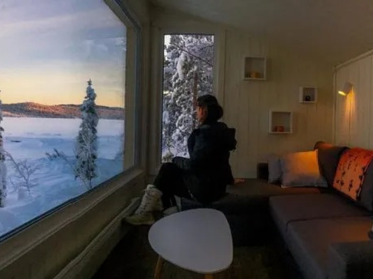 Person seated indoors, observing snowy landscape through large window at sunset.