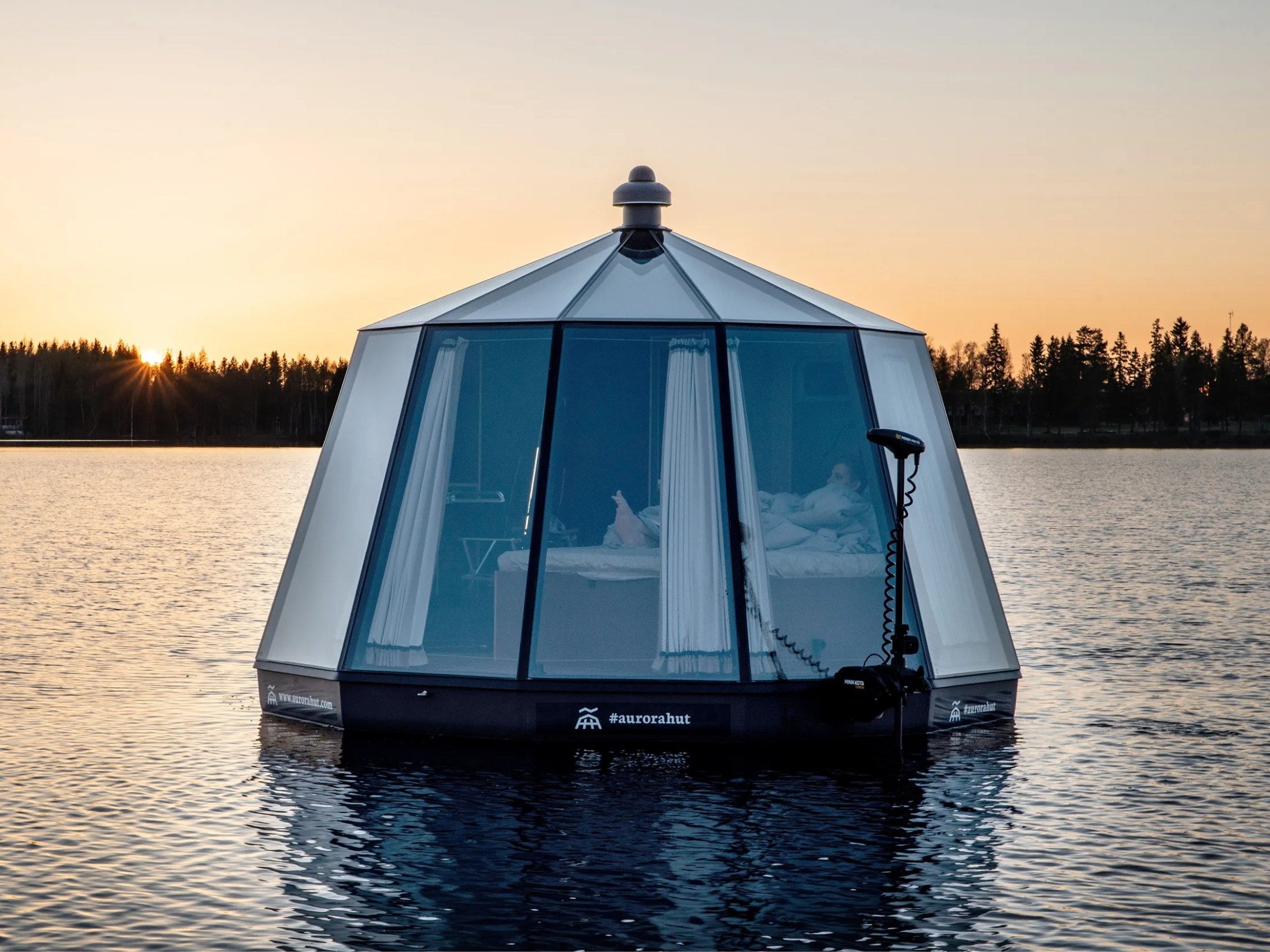 Floating cabin on a lake at sunset, surrounded by forest.