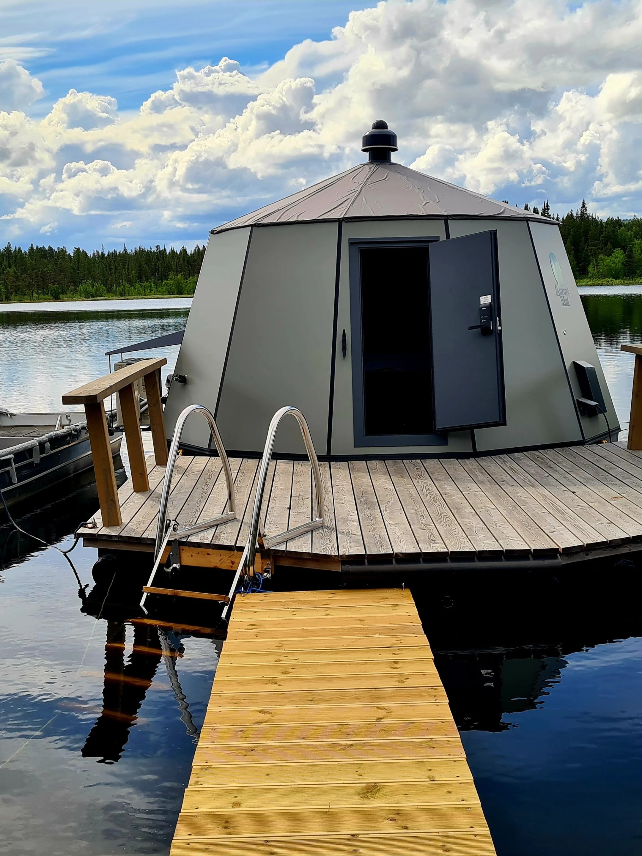 Floating sauna on a lake with wooden deck and boat nearby under a cloudy sky.