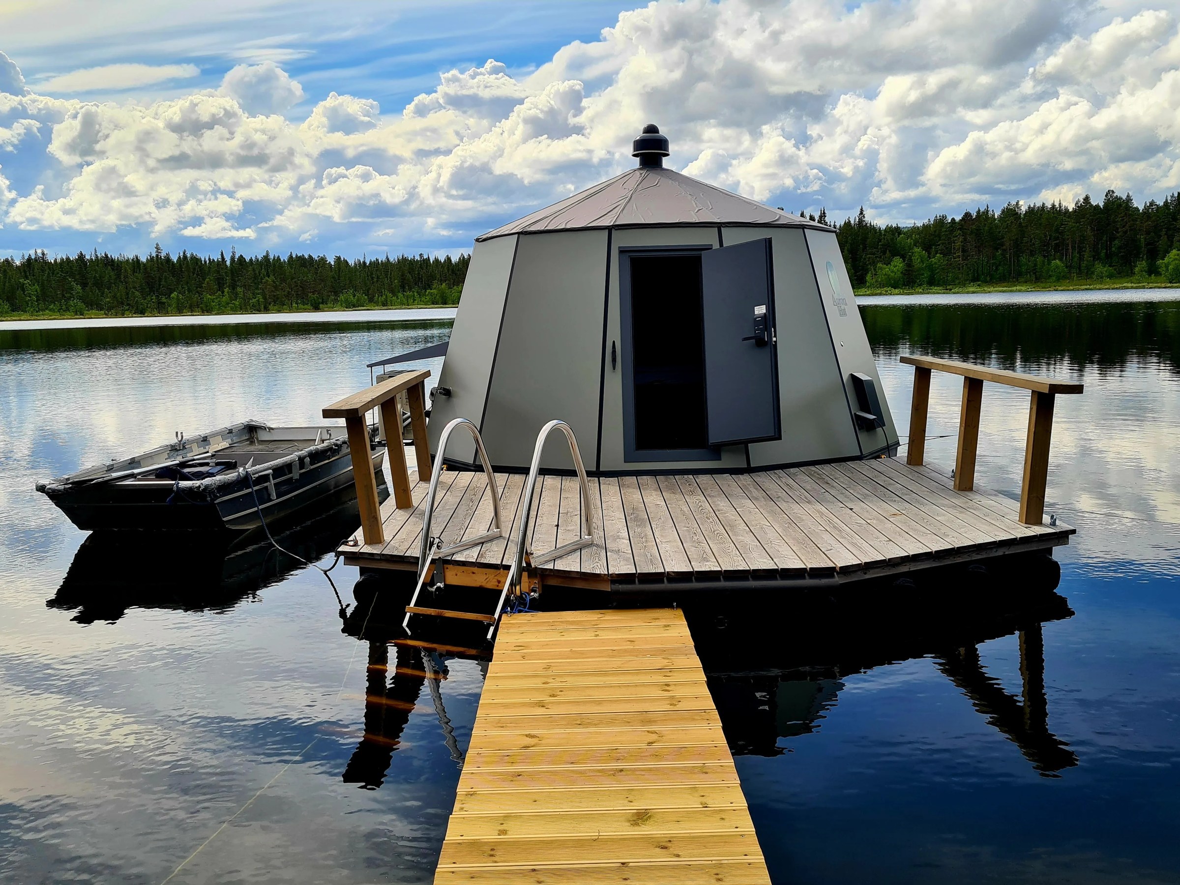 Floating sauna on a lake with wooden deck and boat nearby under a cloudy sky.
