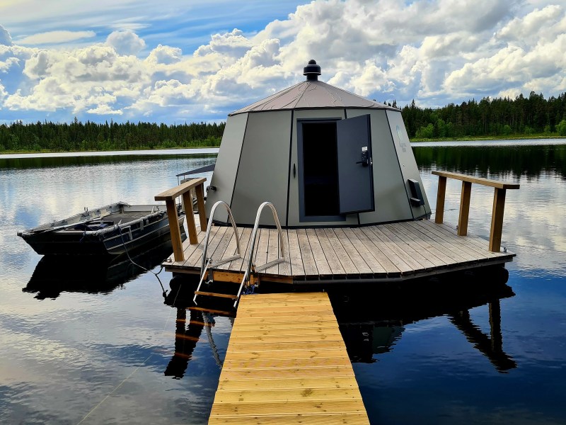 Floating sauna on a lake with wooden deck and boat nearby under a cloudy sky.