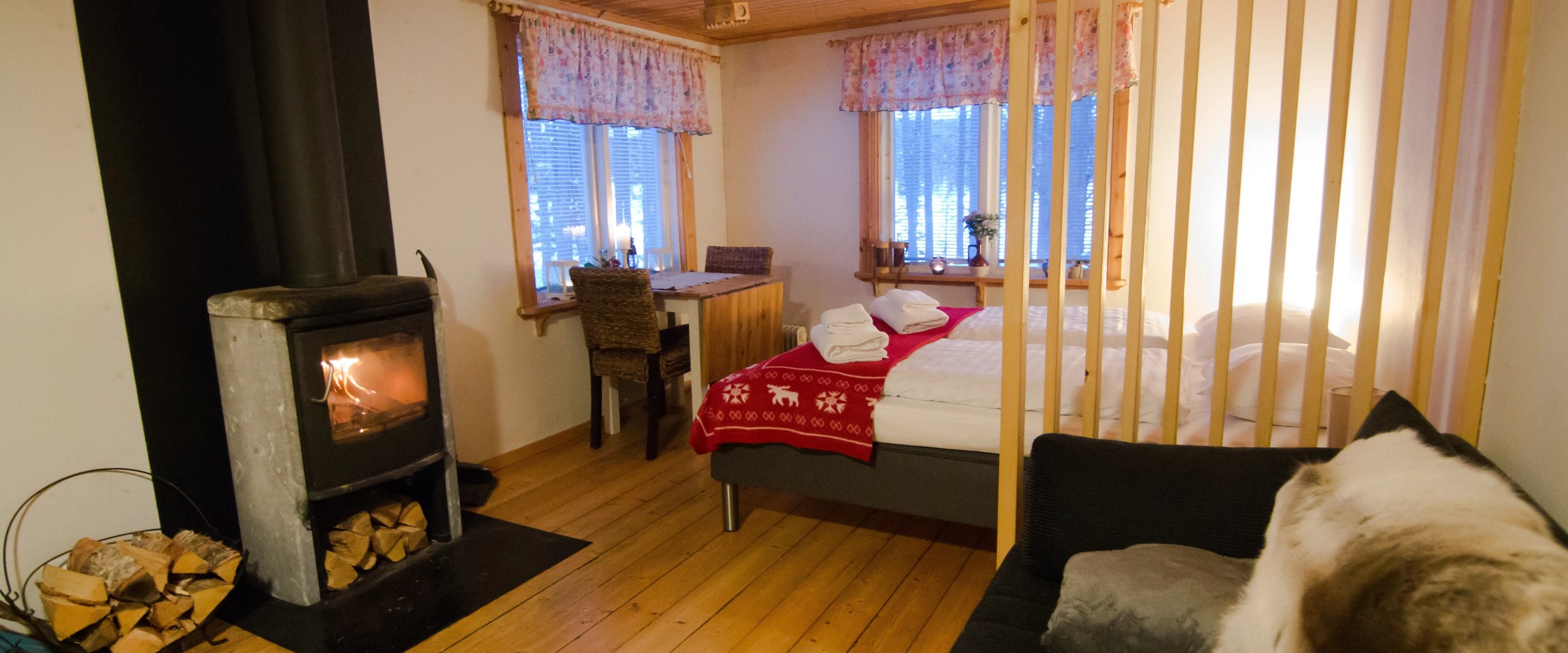 Cozy bedroom with fireplace, wood floors, bed with red blanket, and stacked firewood.