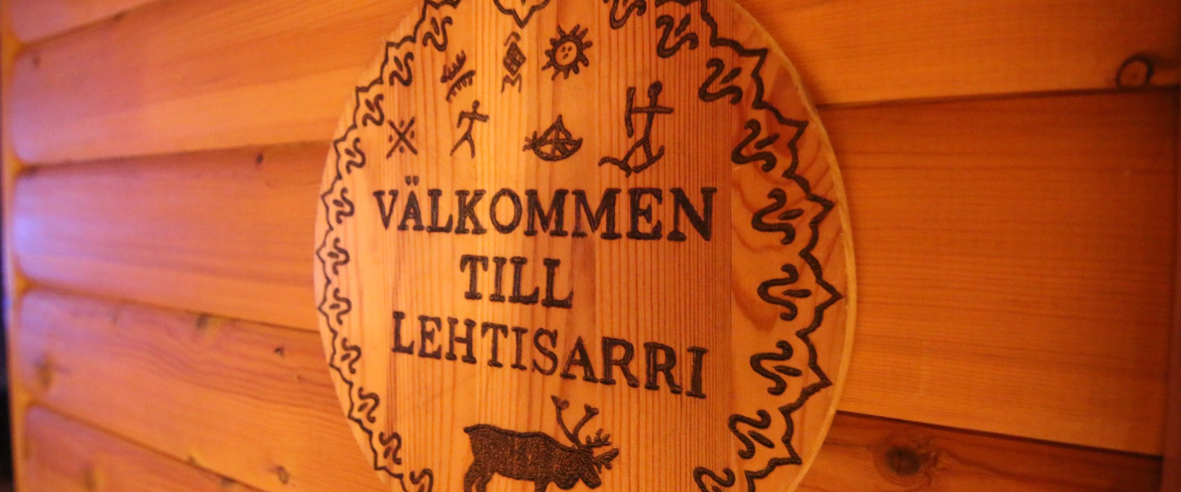 Wooden sign with 'VÄLKOMMEN TILL LEHTISARRI' and carvings on a wooden wall.