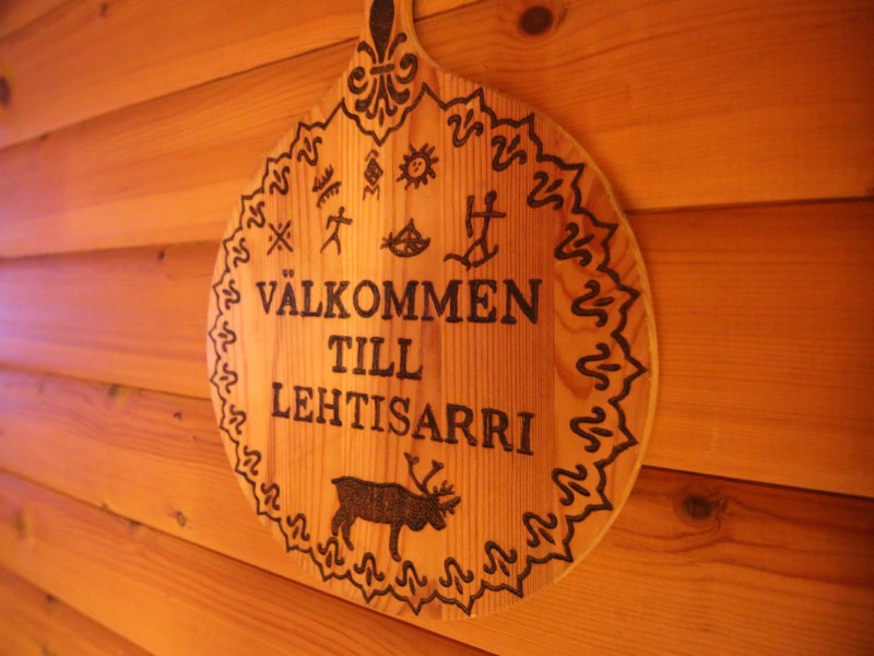 Wooden sign with 'VÄLKOMMEN TILL LEHTISARRI' and carvings on a wooden wall.