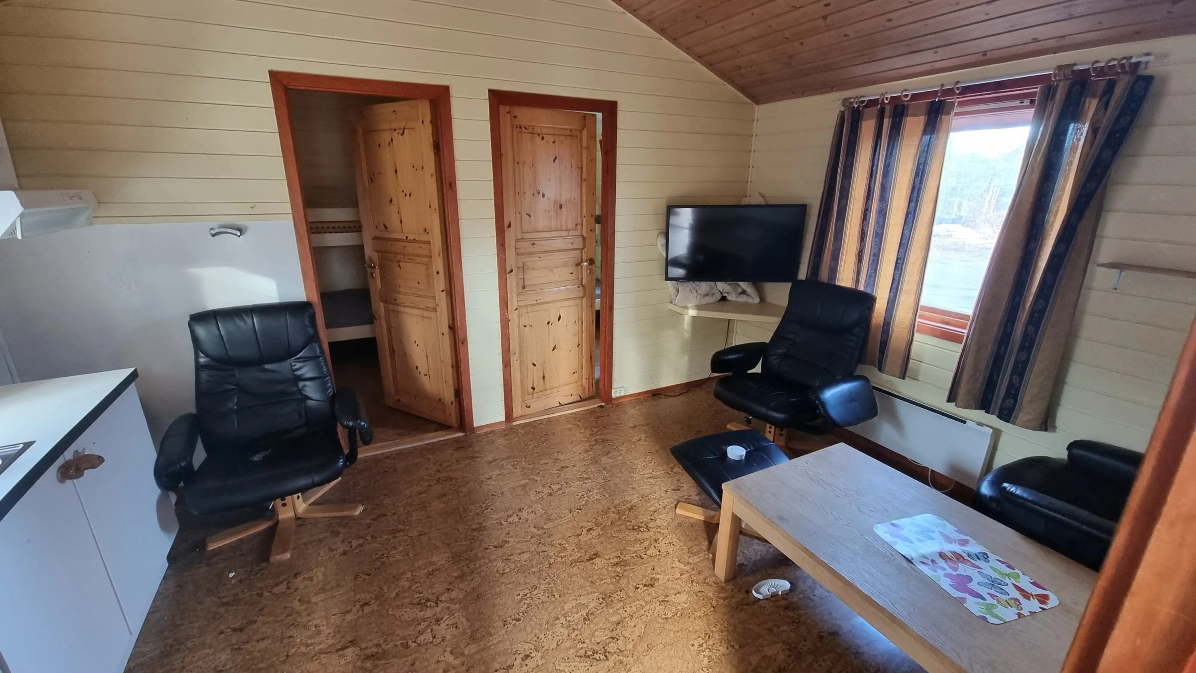 Cozy cabin interior with wooden walls, two black chairs, a TV, table, and striped curtains near a window.