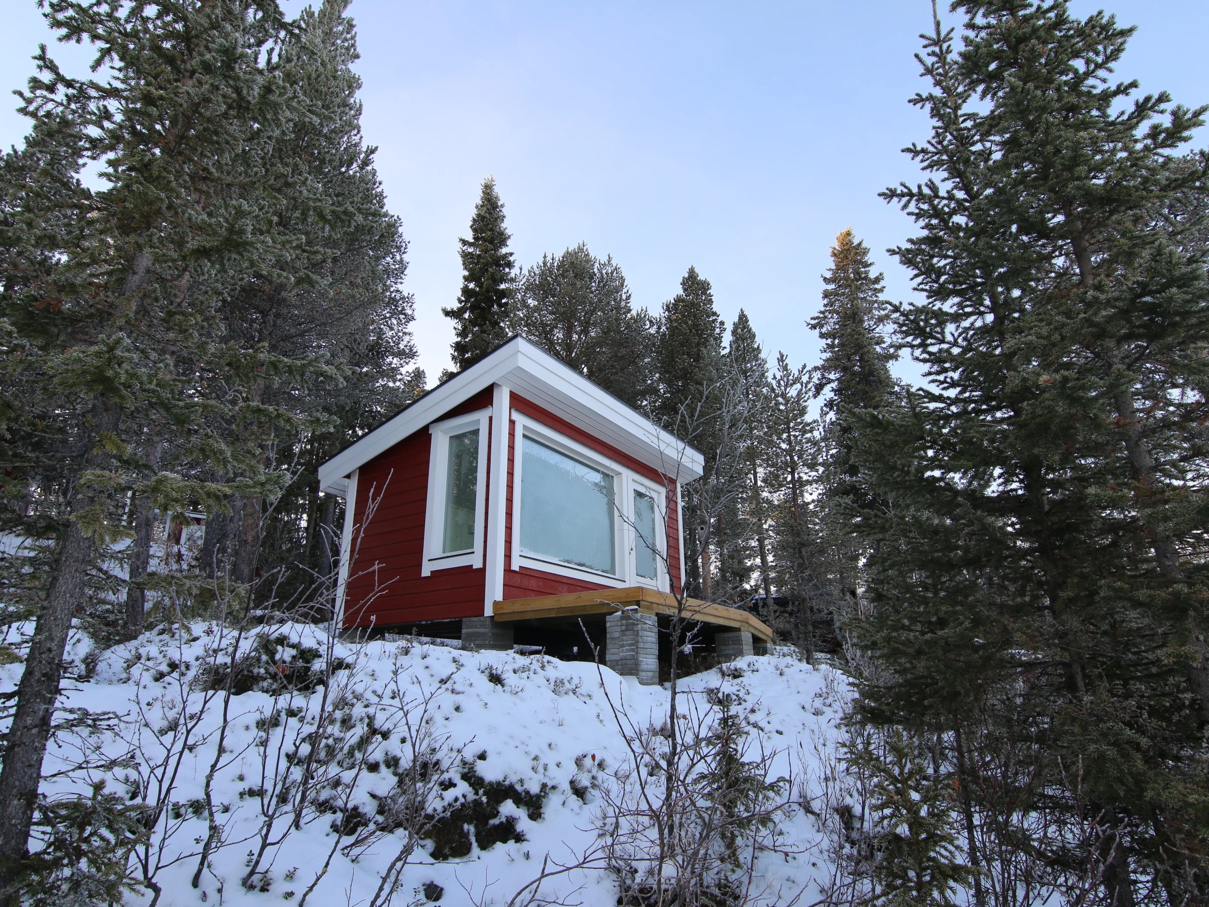 Small red cabin on snowy hillside surrounded by pine trees.