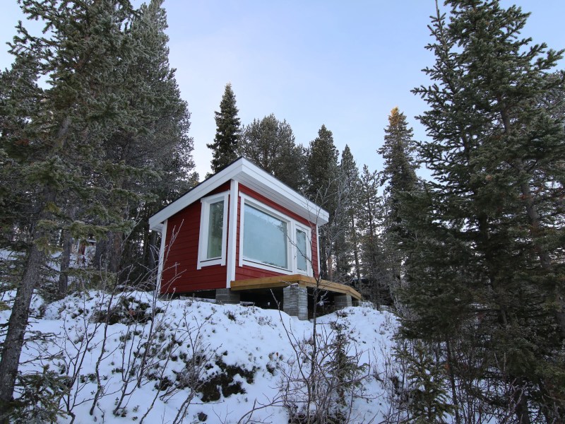 Small red cabin on snowy hillside surrounded by pine trees.