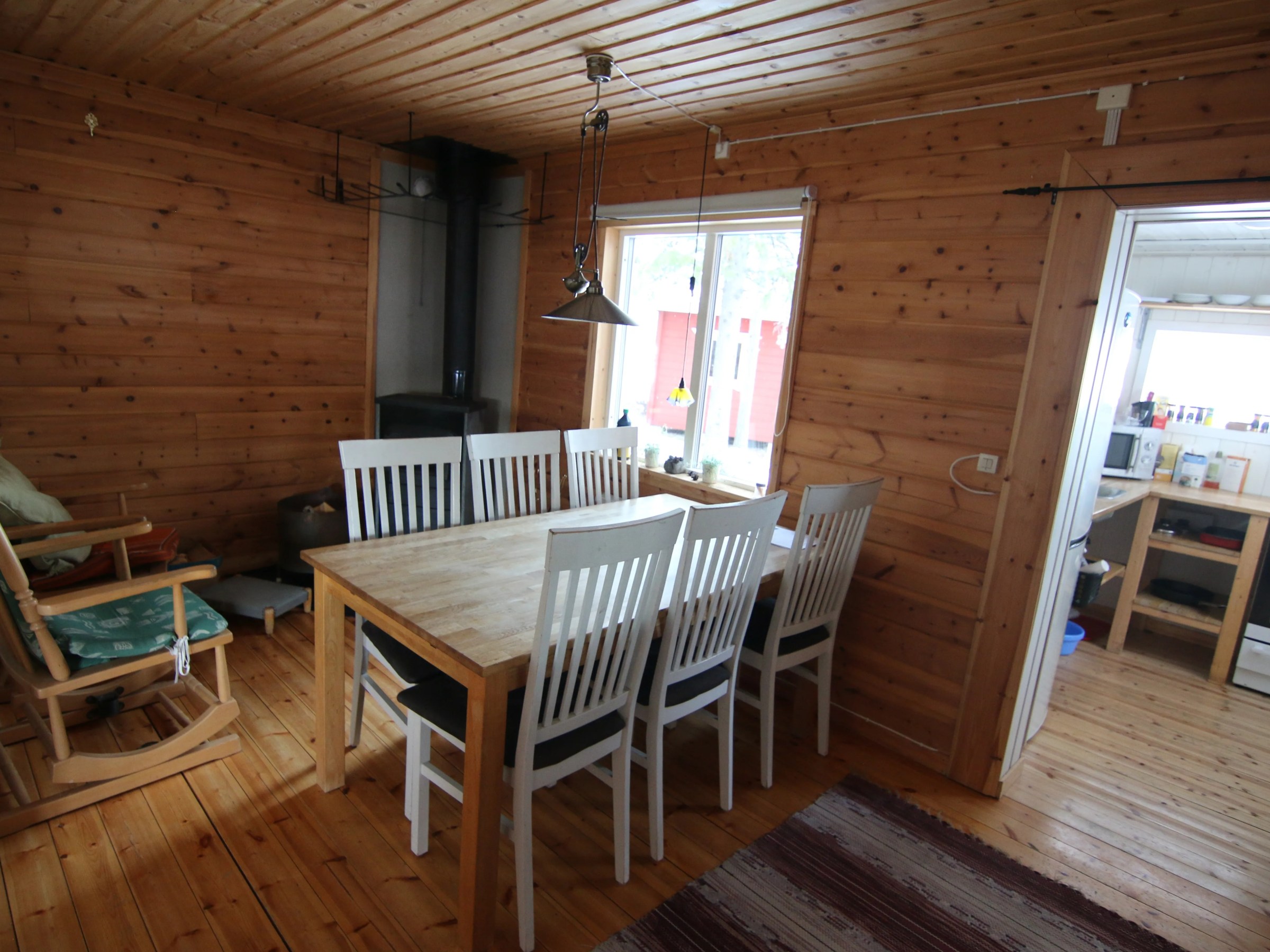 Wood-paneled dining area with table, chairs, rocking chair, and window view of adjacent kitchen.