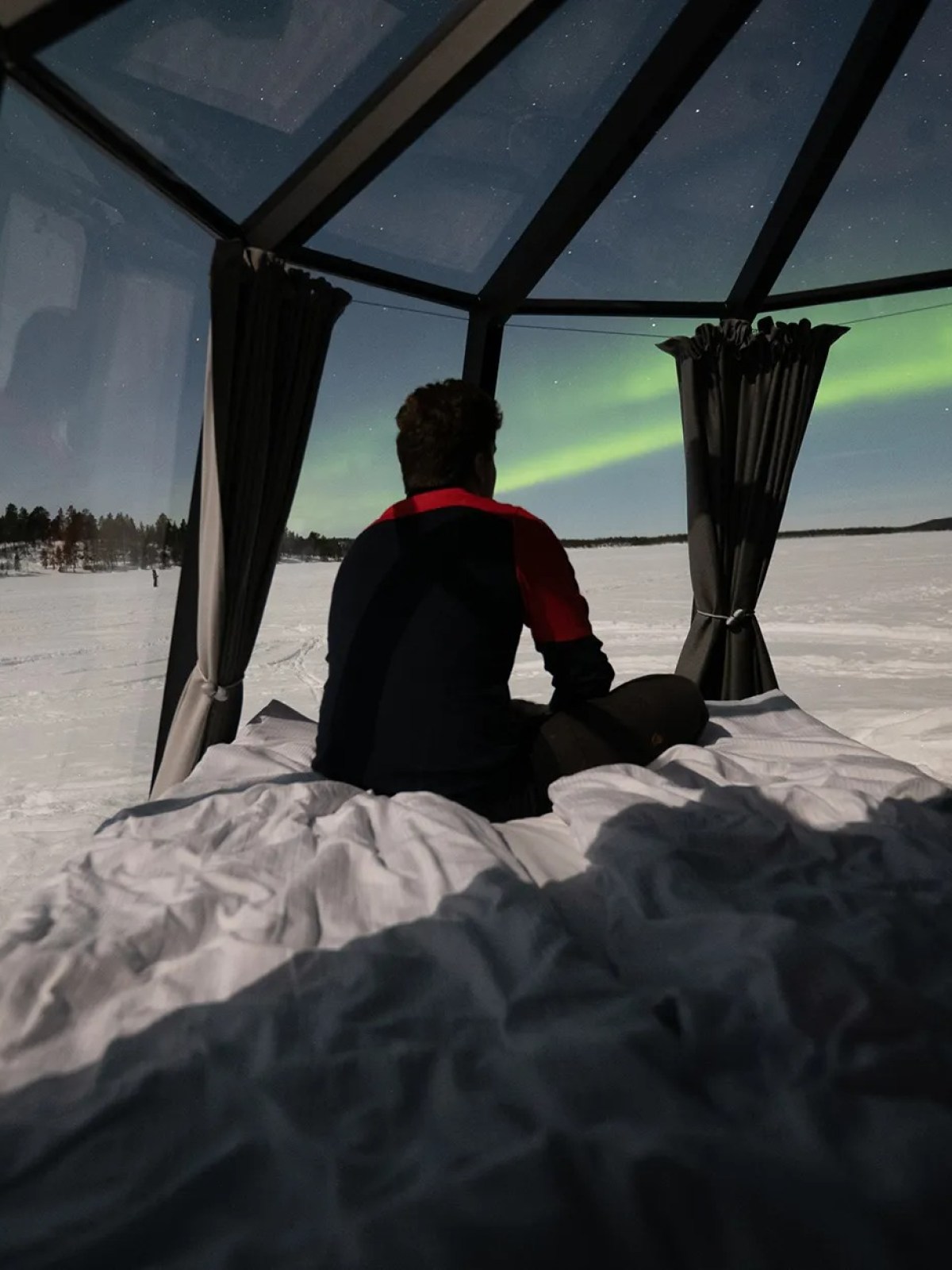Person sitting on bed in glass cabin watching aurora borealis over snowy landscape.
