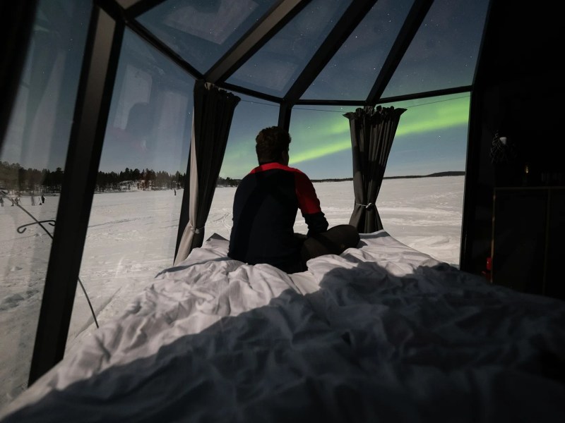 Person sitting on bed in glass cabin watching aurora borealis over snowy landscape.