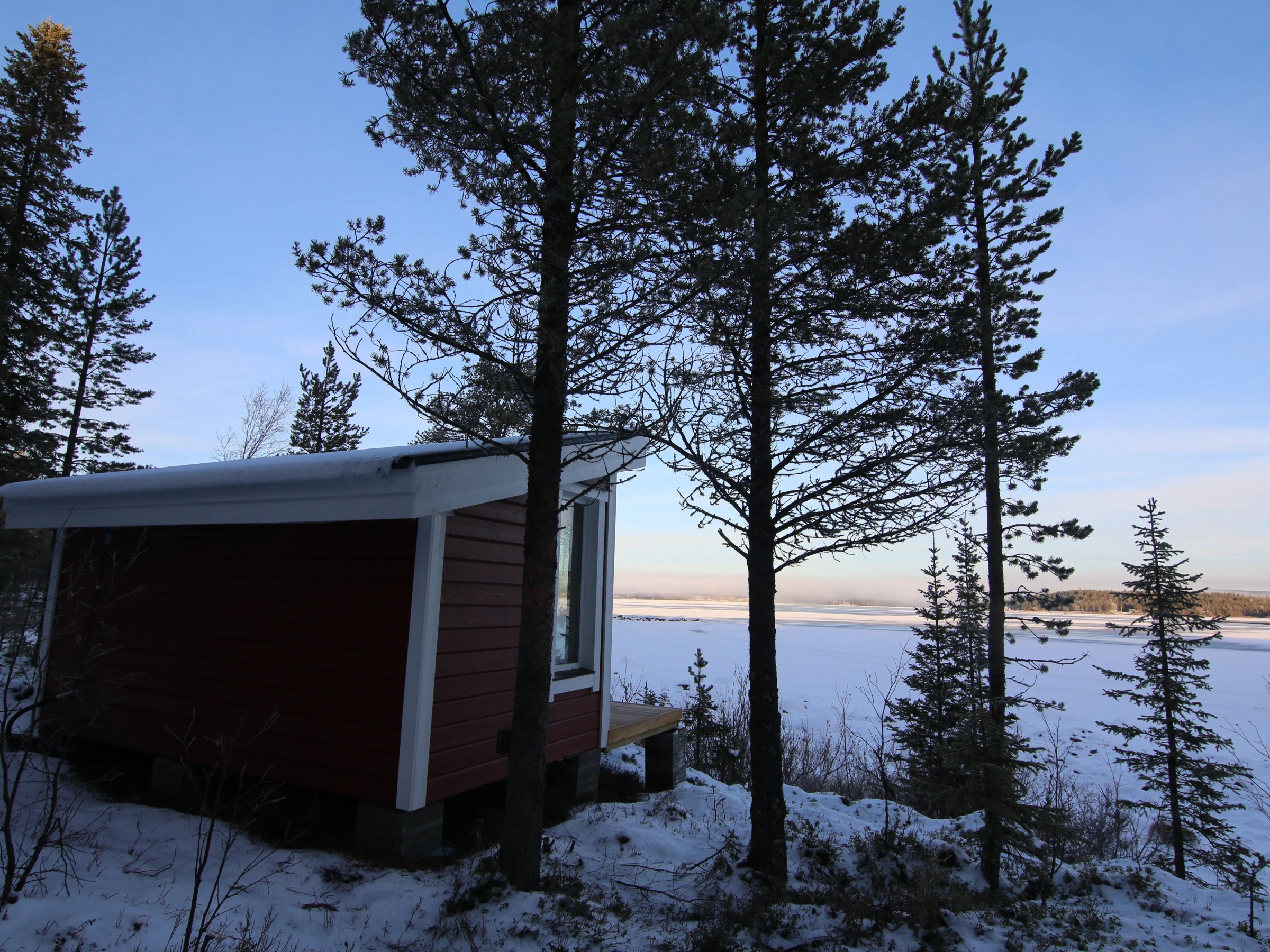 Red cabin in snowy forest overlooking frozen lake under clear sky.