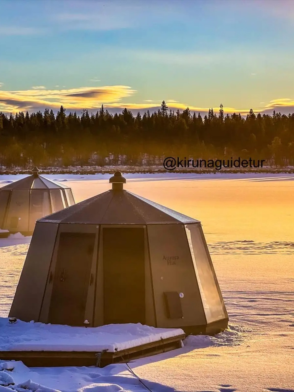 Two octagonal huts on a snowy frozen lake with a sunset and forest background.