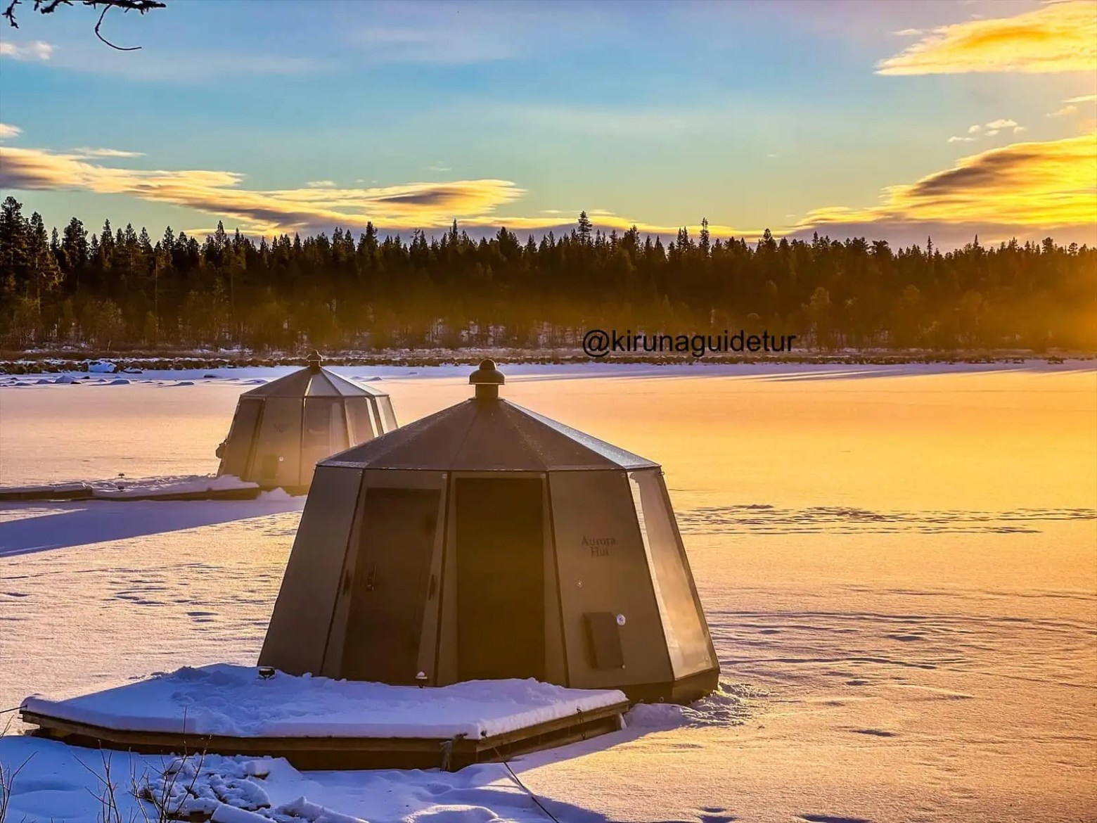 Two octagonal huts on a snowy frozen lake with a sunset and forest background.