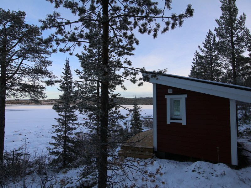 Red cabin beside snowy pine forest and frozen lake under clear sky.