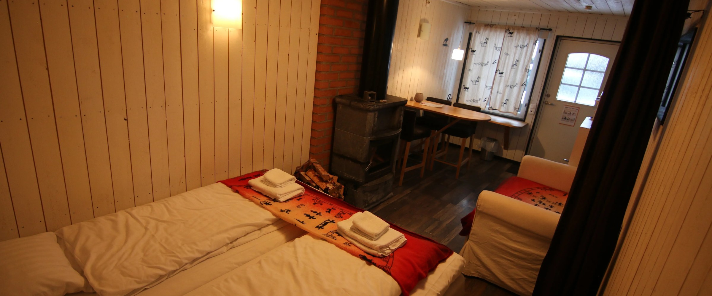 Cozy room with twin beds, a wood stove, and a small dining area near a window.