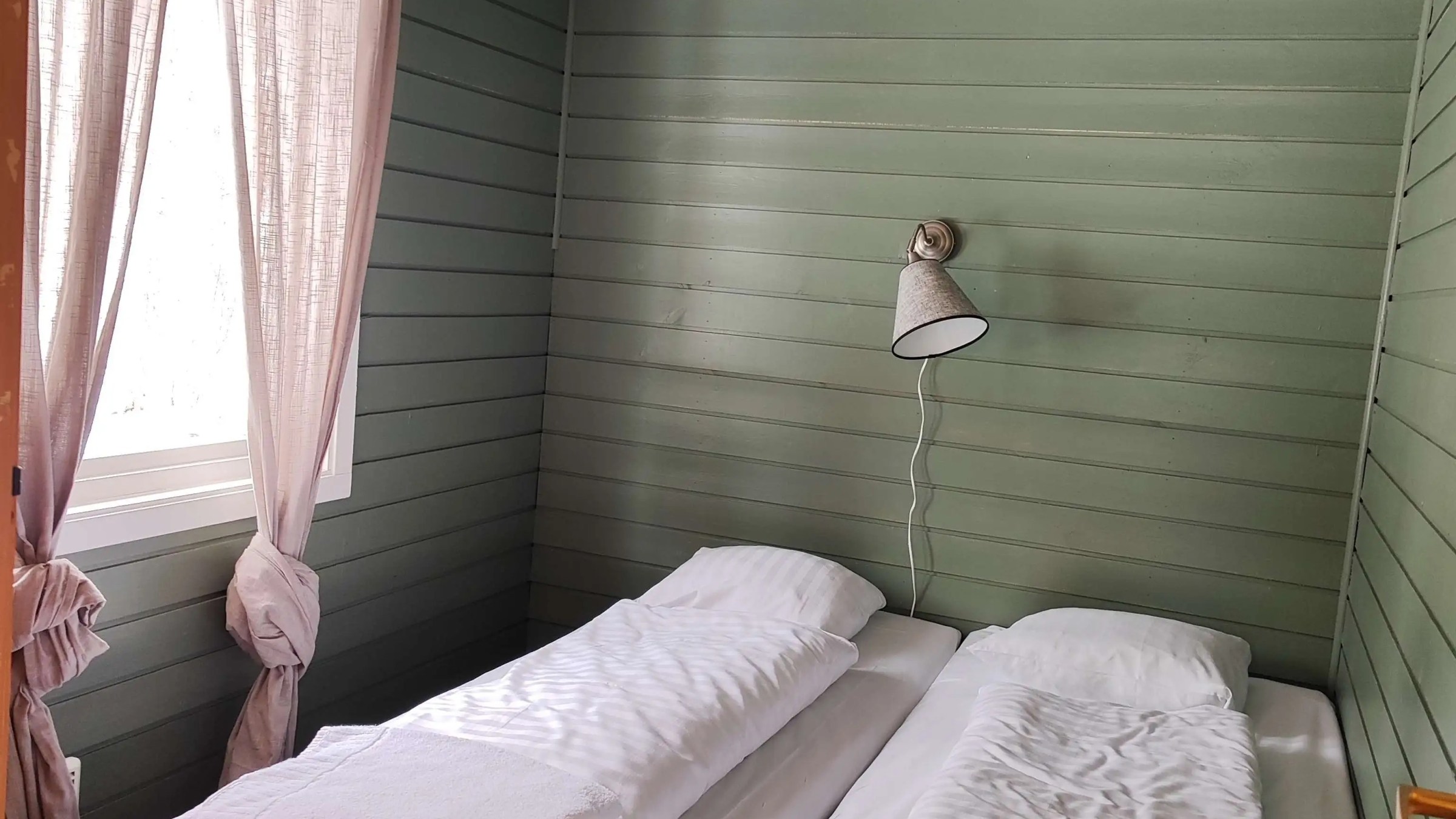 Two neatly made beds in a small room with green wooden walls and a window with curtains.