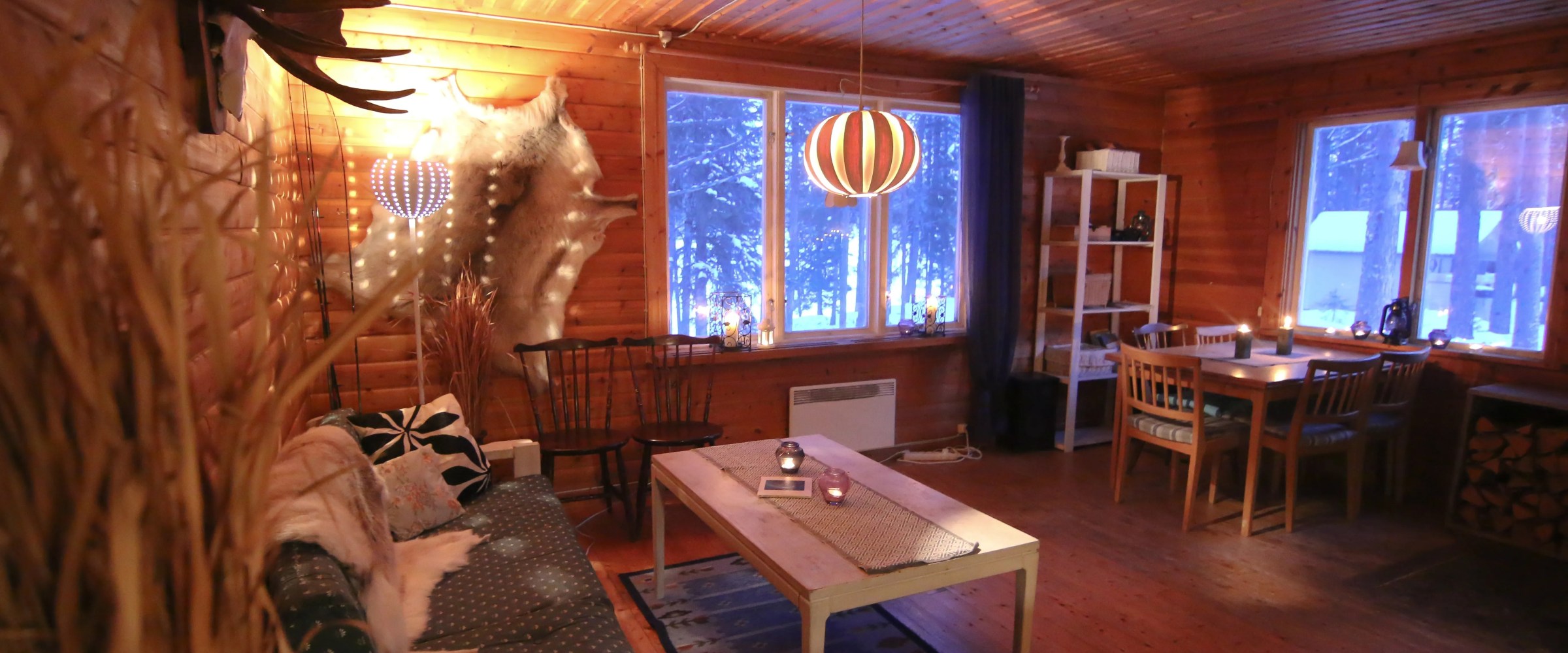 Cozy wooden cabin interior with sofa, table, dining area, and warm lighting.