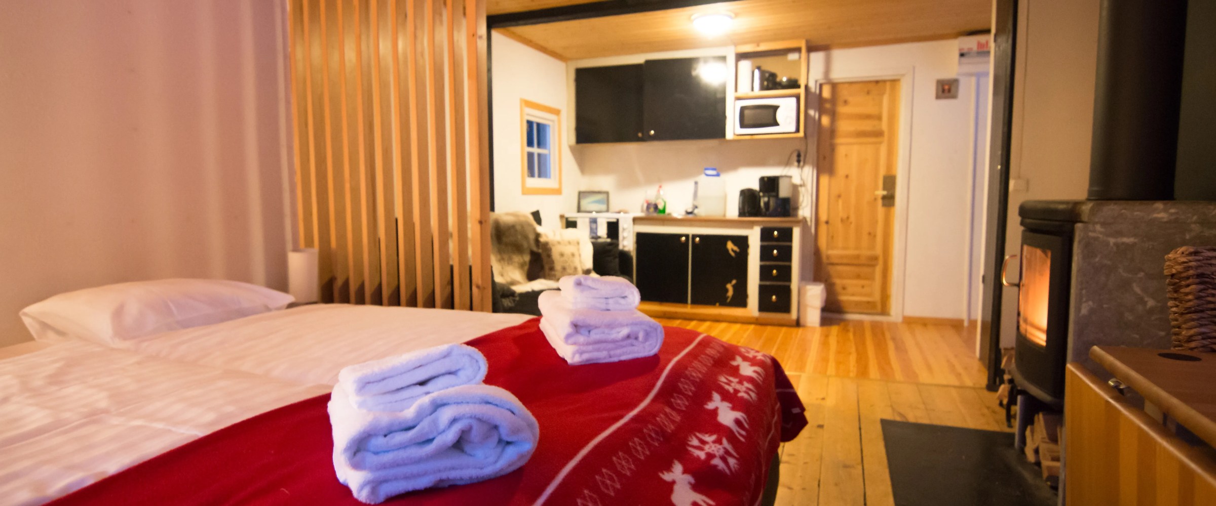 Cozy cabin interior with bed, red blanket, kitchenette, wooden floors, and a wood stove.