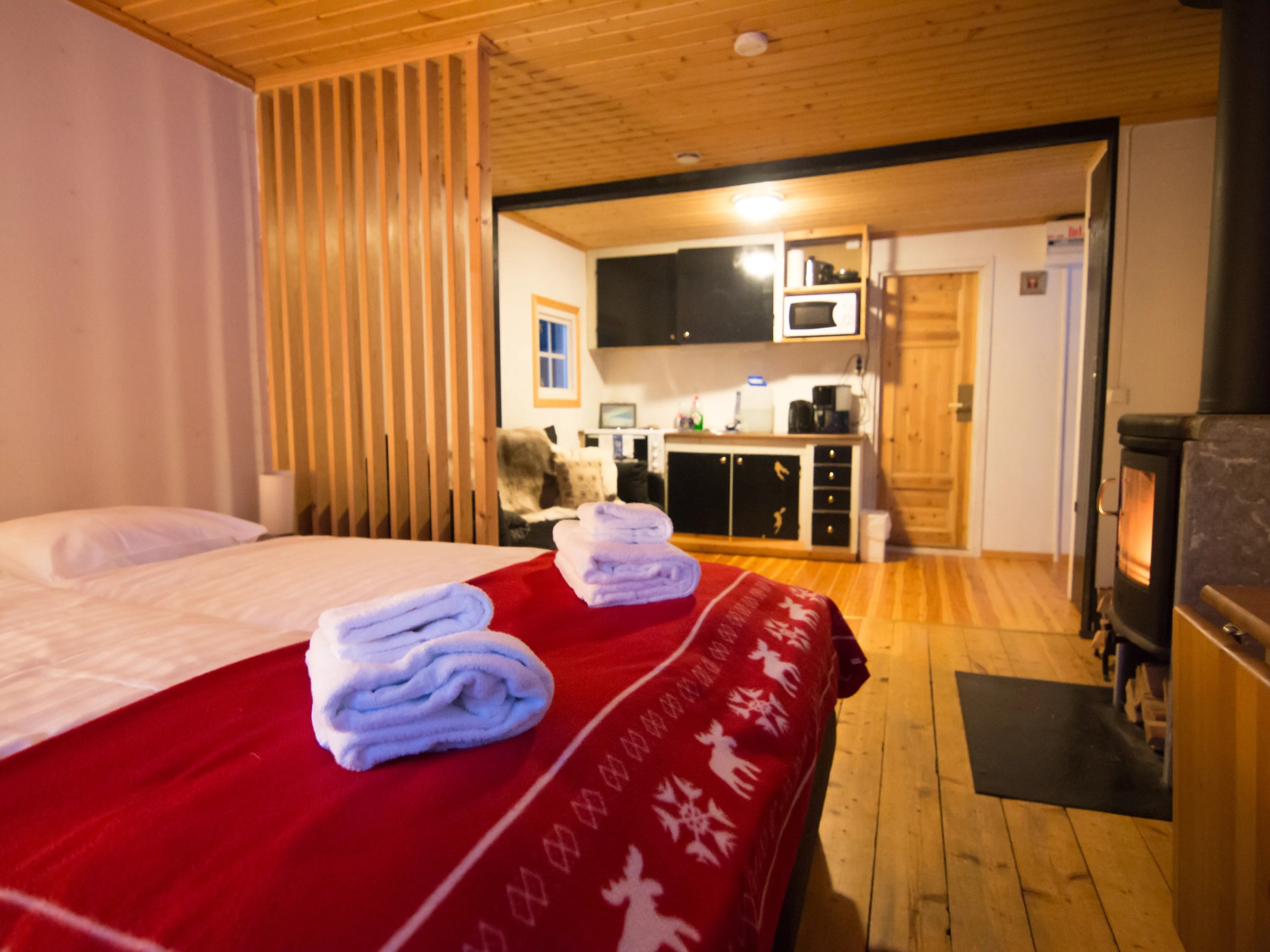 Cozy cabin interior with bed, red blanket, kitchenette, wooden floors, and a wood stove.