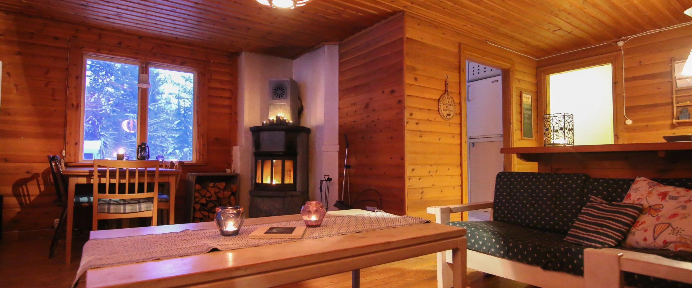 Cozy wooden interior with sofas, table, dining area, and fireplace, lit by warm lighting.